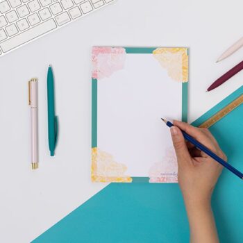 A Floral A5 notepad, with teal border, pink and yellow flowers at the corners. It is on a white desk with a teal corner, and hand is to its right holding a pencil to the page.