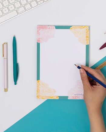 A Floral A5 notepad, with teal border, pink and yellow flowers at the corners. It is on a white desk with a teal corner, and hand is to its right holding a pencil to the page.
