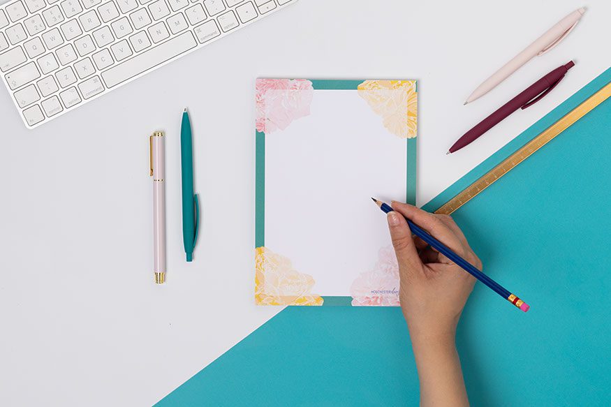 A Floral A5 notepad, with teal border, pink and yellow flowers at the corners. It is on a white desk with a teal corner, and hand is to its right holding a pencil to the page.