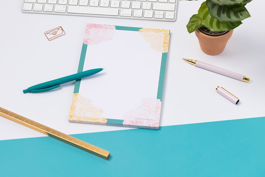 A Floral A5 notepad, with teal border, pink and yellow flowers at the corners. It is on a white desk with a teal corner.