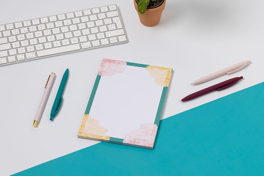 A Floral A5 notepad, with teal border, pink and yellow flowers at the corners. It is on a white desk with a teal corner.