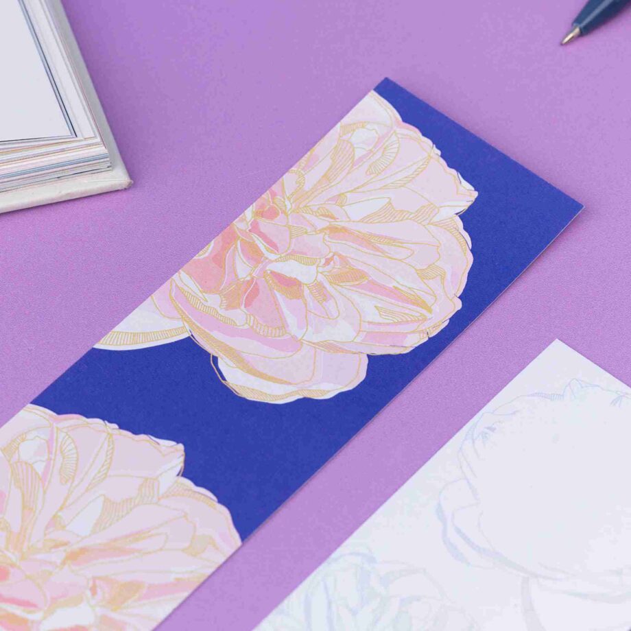 Close up of the Spring Forward Bookmark with a blue background and pink dahlia illustrations and golden yellow outlines. Next to is is an out of focus corner of the bookmark's other side. They are on a lilac desk with an open book in the top left corner.