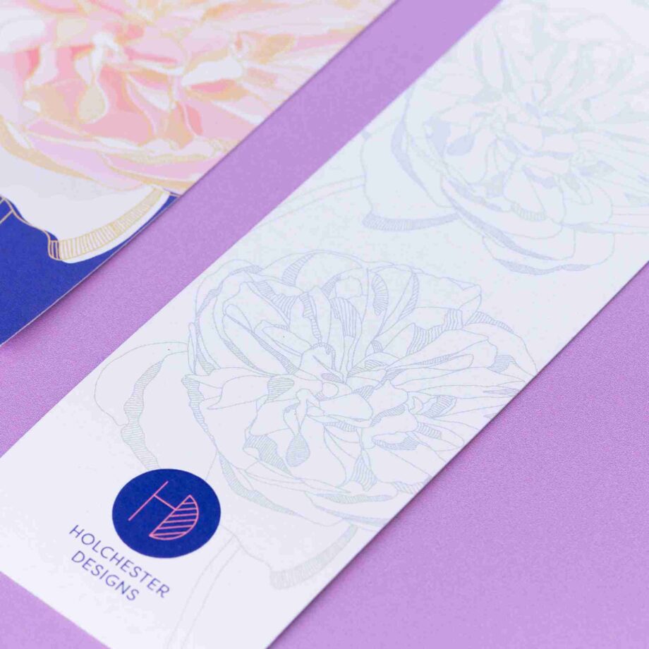 Two bookmarks, showing each side of the Spring Forward Bookmark. In focus is the bottom of the monochrome side, showing the line-drawn motifs in light grey with the Holchester Designs logo. To its left is the full-colour side of the design.