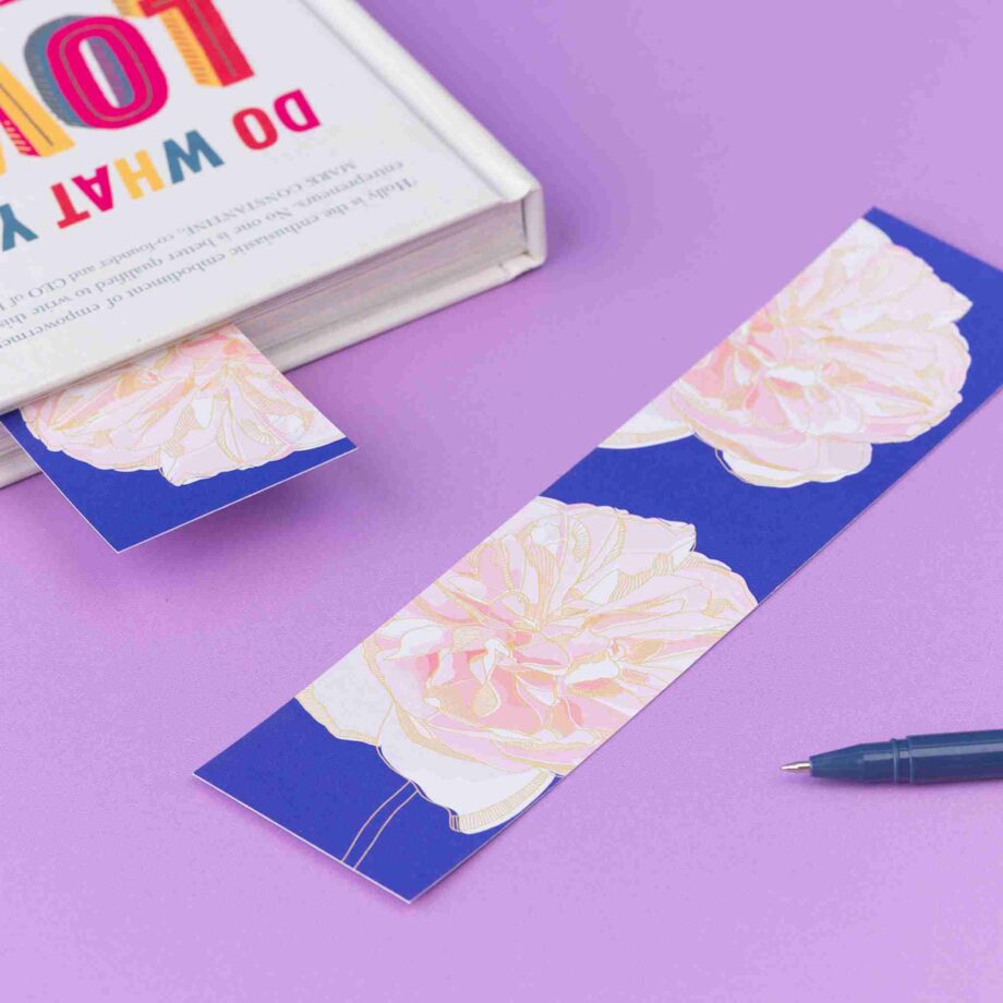 Spring Forward double-sided bookmark is lying, full colour pattern up, on a lilac desk. To its left is a closed book with the same side of the bookmark sticking out of it.