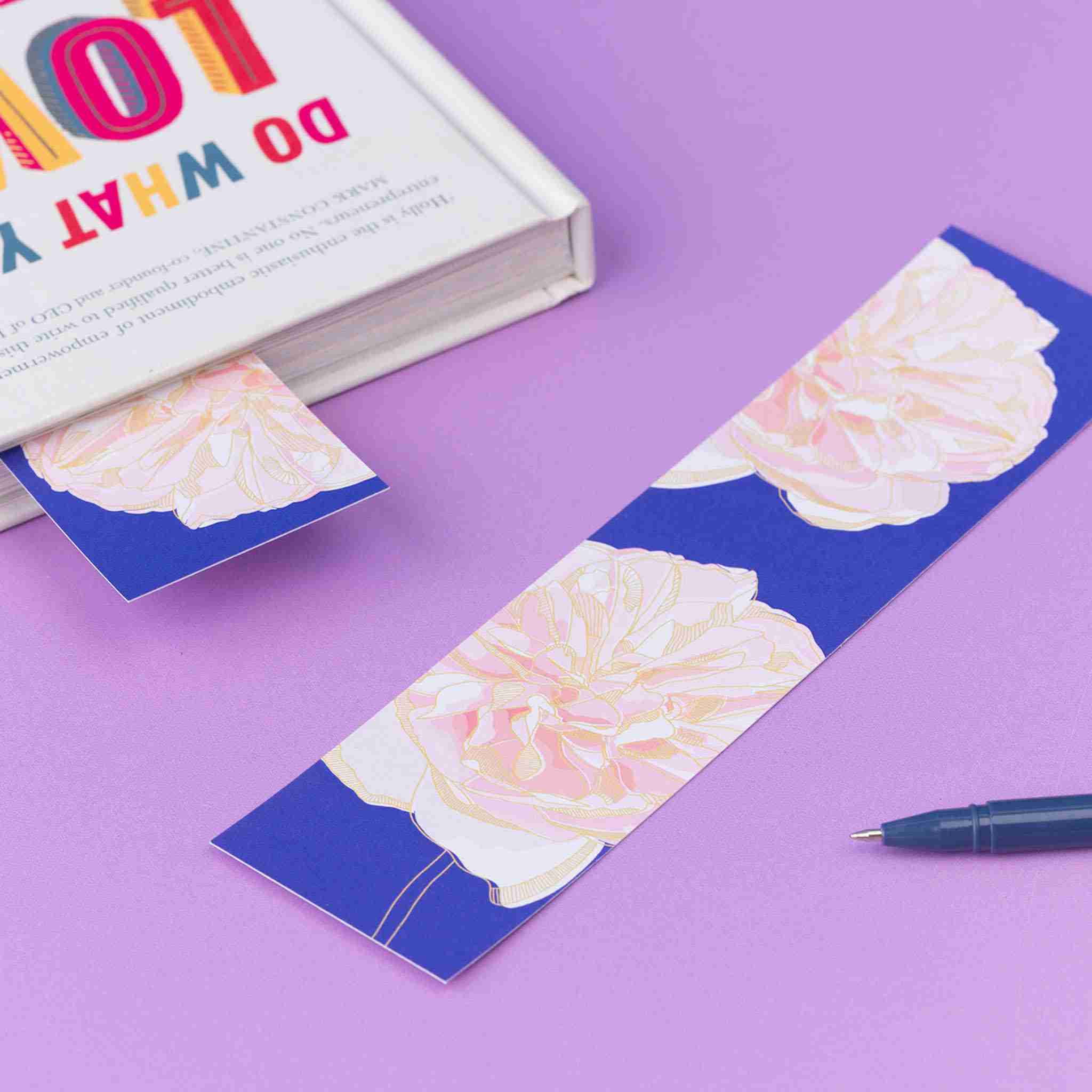 Spring Forward double-sided bookmark is lying, full colour pattern up, on a lilac desk. To its left is a closed book with the same side of the bookmark sticking out of it.