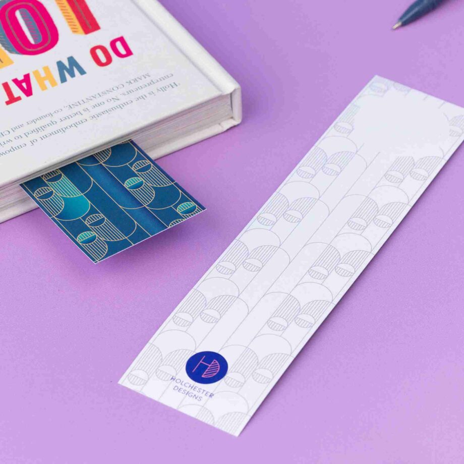 Image of 2 Deco Delights Double sided bookmarks. One on the table has the monochorme side facing up, whilst the other has the full colour side facing up and is partially in a book to the top right corner.