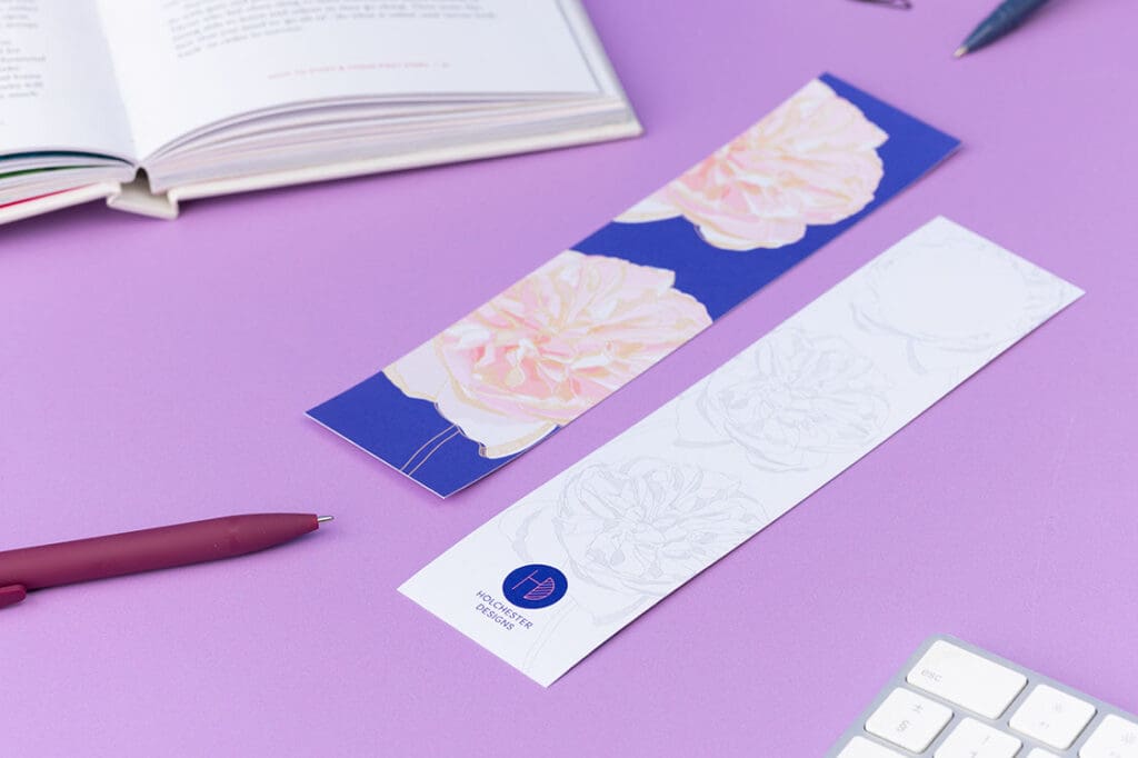 Two bookmarks, showing each side of the Spring Forward Bookmark. One side has a blue background with pink dhalia illustrations and golden ellow outlines, the other side is white with light grey outlines of the dahlia. They are on a lilac desk with an open book, pens and keyboard around them.