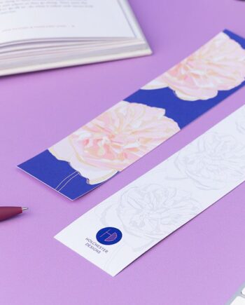 Two bookmarks, showing each side of the Spring Forward Bookmark. One side has a blue background with pink dhalia illustrations and golden ellow outlines, the other side is white with light grey outlines of the dahlia. They are on a lilac desk with an open book, pens and keyboard around them.