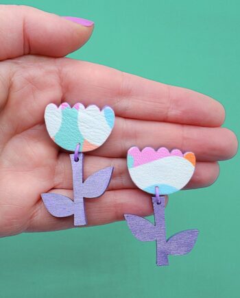 A pair of colourful flower-shaped drop earrings featuring pastel rainbow faux leather petals and metallic purple wooden stems. These playful, statement earrings add a whimsical touch to any outfit.