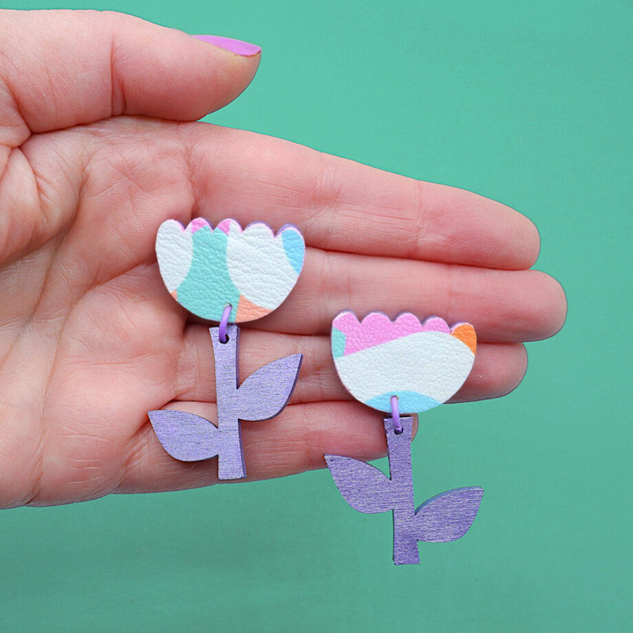 A pair of colourful flower-shaped drop earrings featuring pastel rainbow faux leather petals and metallic purple wooden stems. These playful, statement earrings add a whimsical touch to any outfit.