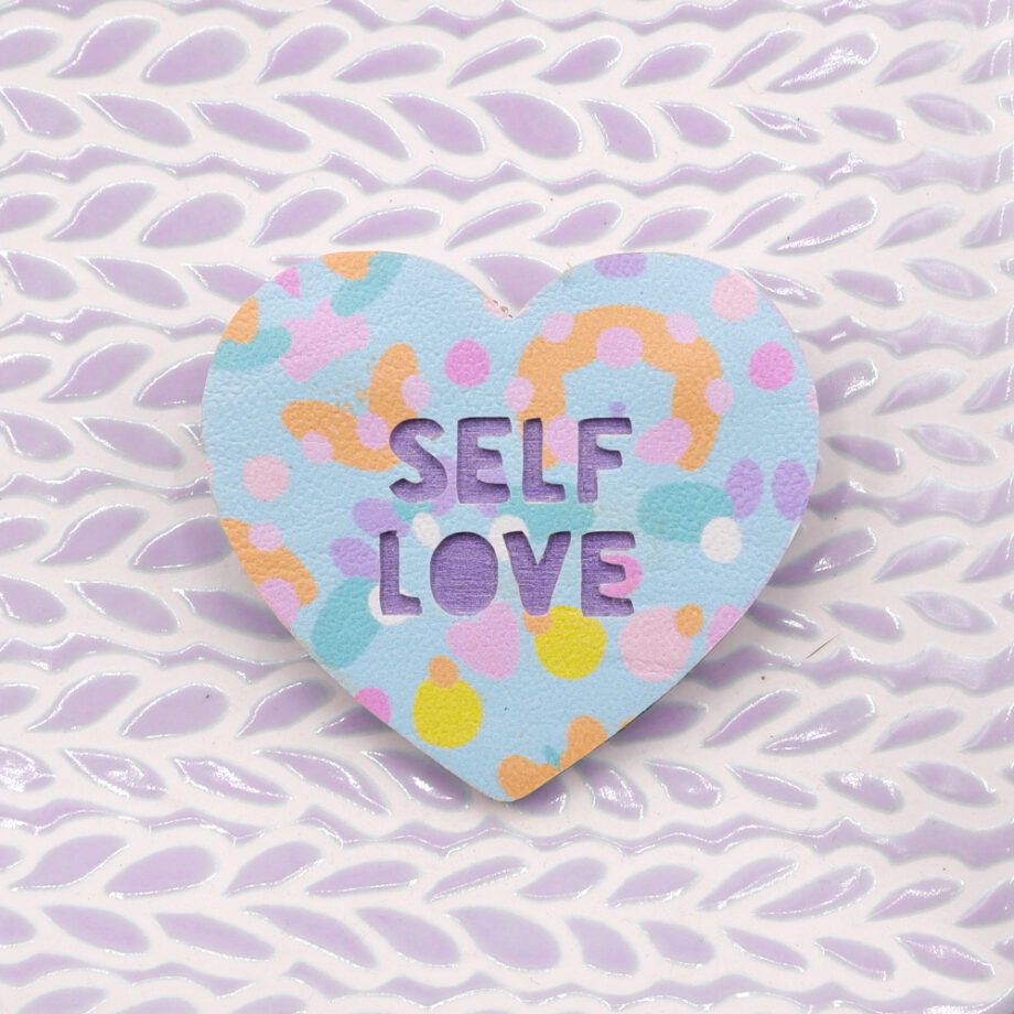 This pretty brooch is reminder to Love Yourself. It's handmade from faux leather printed with an original pastel blue, peach and pink abstract design attached to purple metallic painted plywood with the phrase Self Love cut out from the fabric.