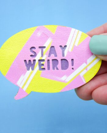 Bright yellow speech bubble brooch with pink graffiti-style strokes and "Stay Weird" cut-out text, backed with metallic purple-painted wood, held in a hand against a blue background.