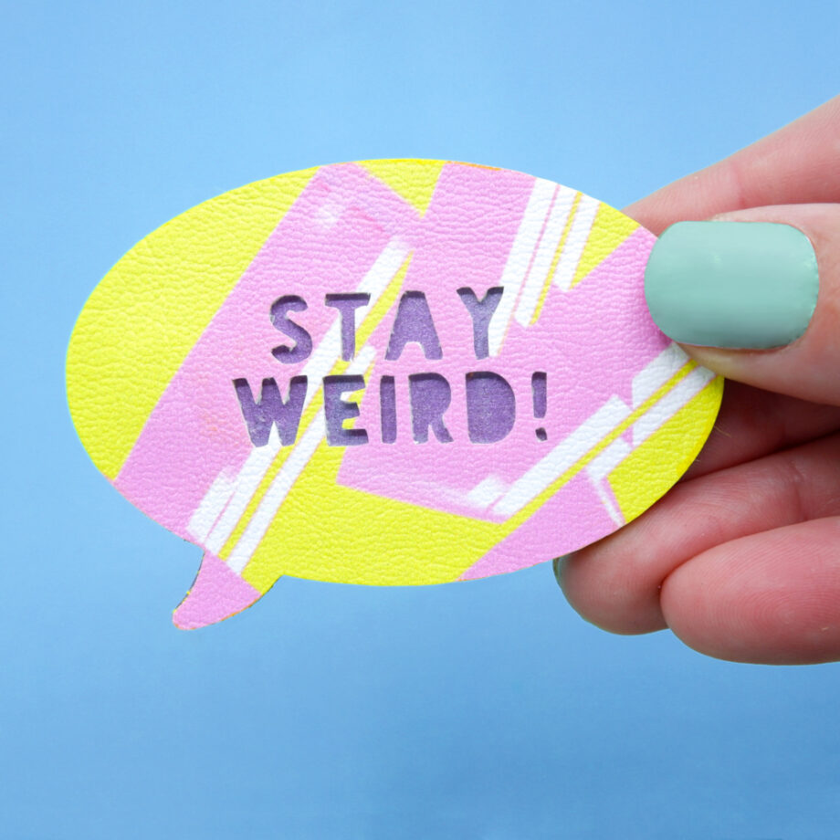 Bright yellow speech bubble brooch with pink graffiti-style strokes and "Stay Weird" cut-out text, backed with metallic purple-painted wood, held in a hand against a blue background.