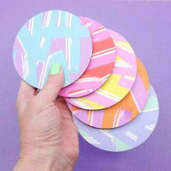 A set of round colourful faux leather coasters with bold abstract patterns in pastel and vibrant hues, displayed against a purple background, whilst being held by a white woman's hand.