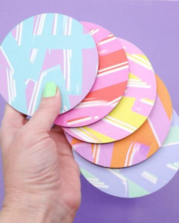 A set of round colourful faux leather coasters with bold abstract patterns in pastel and vibrant hues, displayed against a purple background, whilst being held by a white woman's hand.