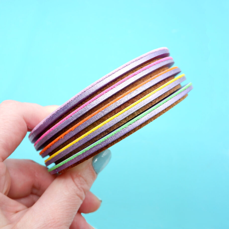 A set of round colourful faux leather coasters with bold abstract patterns in pastel and vibrant hues, displayed against a purple background, whilst being held by a white woman's hand.