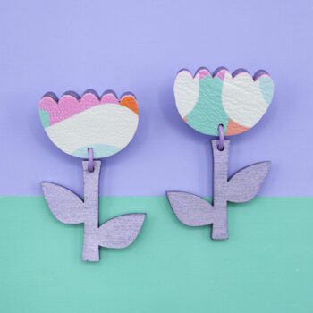 Pastel floral dangle earrings with faux leather tulip tops and lilac-painted wooden stems. The layered paper background creates the illusion of flowers growing in a garden, with green as the grass and soft blue as the sky. These lightweight statement earrings capture the charm of spring with playful colours and a whimsical design.