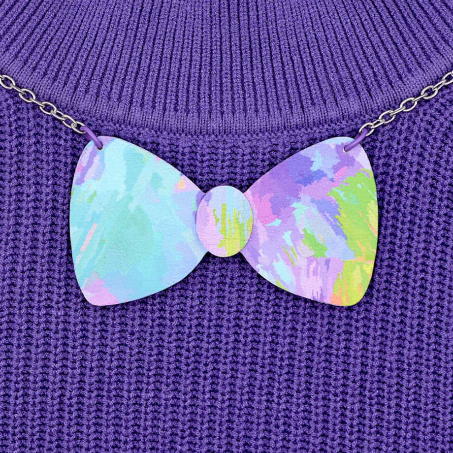 A pastel bow statement necklace with an abstract painterly design, featuring soft washes of purple, blue turquoise and pink. Fabric print is designed by me and printed on to faux leather. Shown worn against a purple knit jumper.