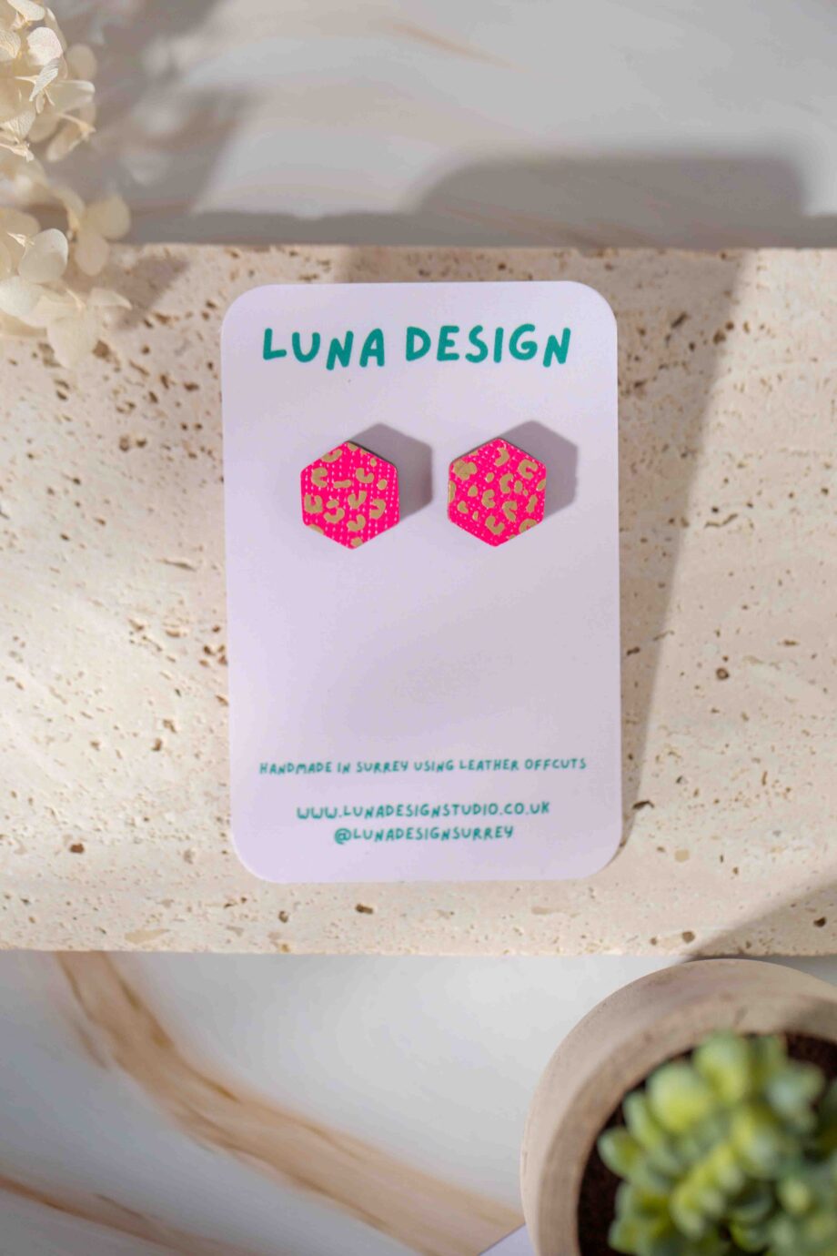 A pair of hexagonal stud earrings with a vibrant neon pink base and gold leopard print detailing, displayed on a white Luna Design card. The card features green branding text and mentions that the earrings are handmade in Surrey using leather offcuts. The earrings are photographed on a textured stone surface with soft natural lighting, accompanied by dried flowers and a small succulent plant in the background.