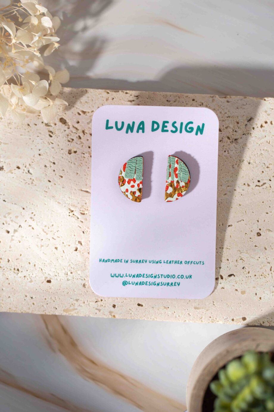 A pair of semi-circle stud earrings with a hand-painted abstract design in green, white, and gold with red accents, displayed on a Luna Design branded card against a neutral stone background.