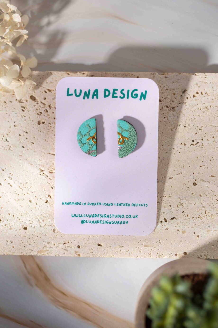 A pair of semi-circle stud earrings made from upcycled leather, featuring a textured turquoise surface with delicate gold detailing. Displayed on a "Luna Design" branded card, with a natural stone background and soft lighting.