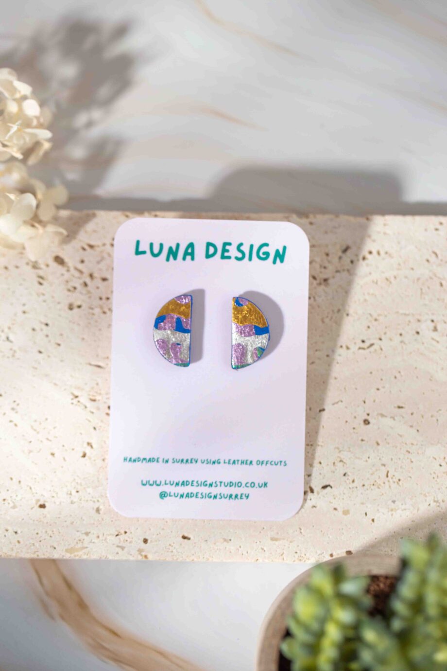 A pair of handmade semi-circle stud earrings crafted from upcycled leather with a colorful abstract pattern featuring gold, blue, lilac, and white tones. Displayed on a Luna Design card.