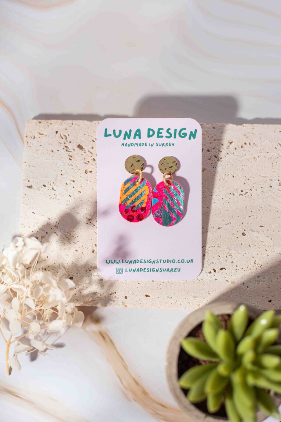 A pair of gold plated stud drop earrings with a hand-painted abstract design in pink, gold and green accents displayed on a Luna Design branded card against a neutral stone background.