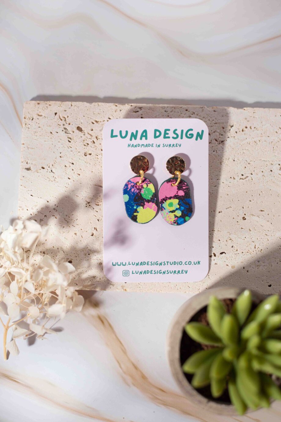 A pair of gold plated stud drop earrings with splatter paint design displayed on a Luna Design branded card against a neutral stone background.