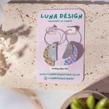 A pair of handmade half-moon leather hoop earrings from Luna Design, displayed on a branded backing card. The earrings feature a mix of silver and iridescent leather pieces, hanging from 925 sterling silver hoops. The display is styled with natural textures, dried flowers, and a small succulent for a soft, organic feel.