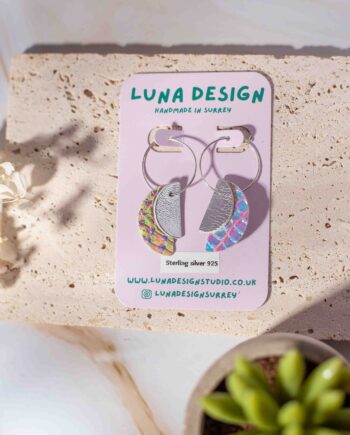 A pair of handmade half-moon leather hoop earrings from Luna Design, displayed on a branded backing card. The earrings feature a mix of silver and iridescent leather pieces, hanging from 925 sterling silver hoops. The display is styled with natural textures, dried flowers, and a small succulent for a soft, organic feel.