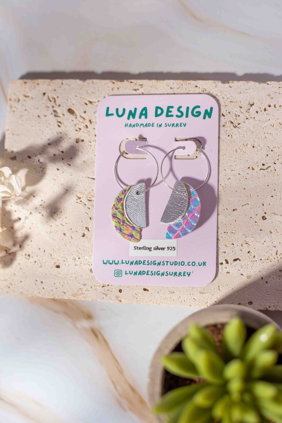 A pair of handmade half-moon leather hoop earrings from Luna Design, displayed on a branded backing card. The earrings feature a mix of silver and iridescent leather pieces, hanging from 925 sterling silver hoops. The display is styled with natural textures, dried flowers, and a small succulent for a soft, organic feel.