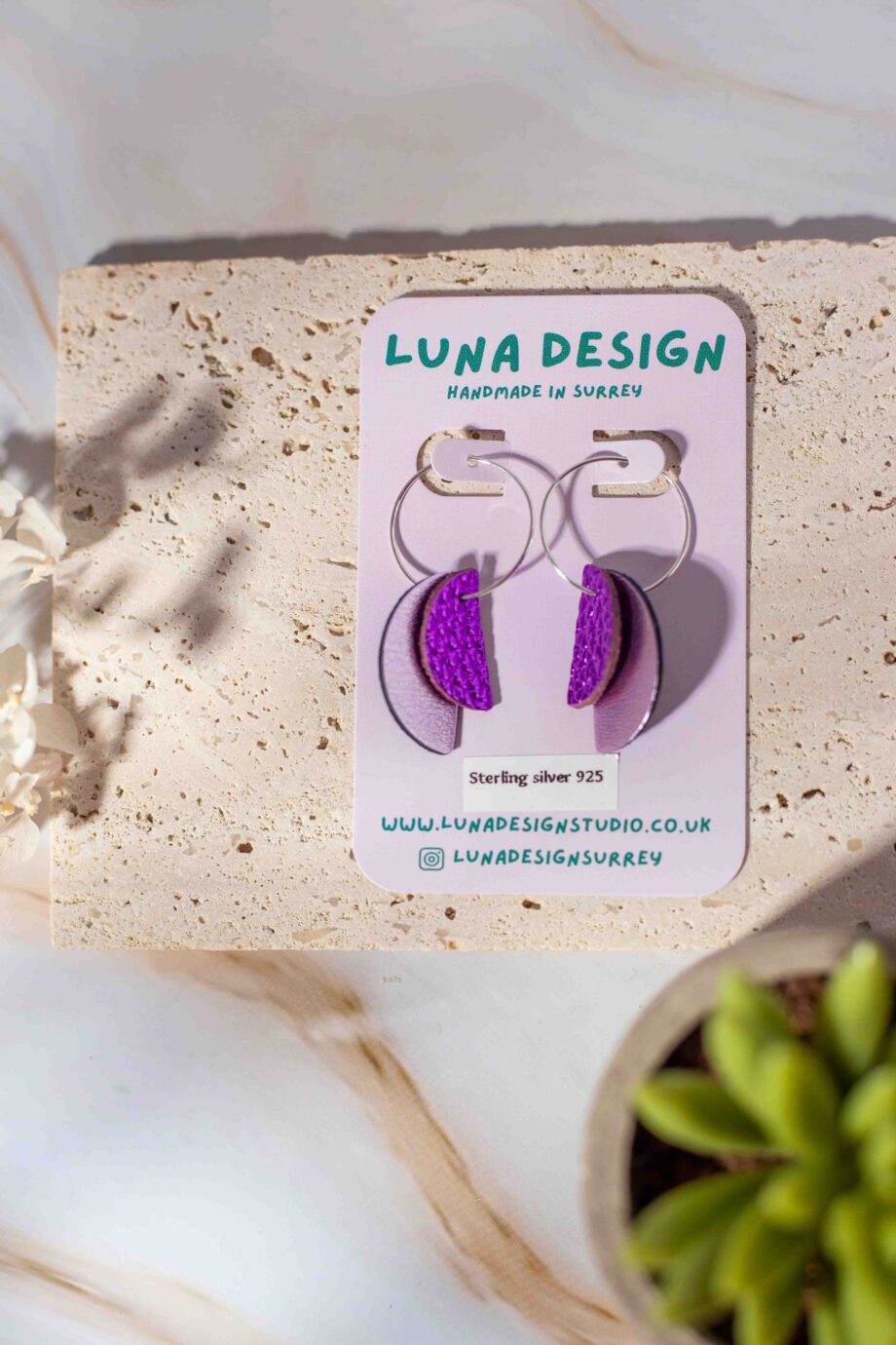 A pair of handmade half-moon leather hoop earrings from Luna Design, displayed on a branded backing card. The earrings feature a mix of purple and pale pink leather pieces, hanging from 925 sterling silver hoops. The display is styled with natural textures, dried flowers, and a small succulent for a soft, organic feel.