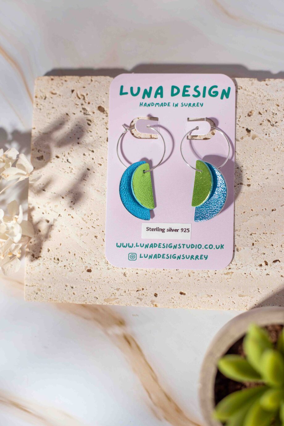 A pair of handmade half-moon leather hoop earrings from Luna Design, displayed on a branded backing card. The earrings feature a mix of blue and green leather pieces, hanging from 925 sterling silver hoops. The display is styled with natural textures, dried flowers, and a small succulent for a soft, organic feel.