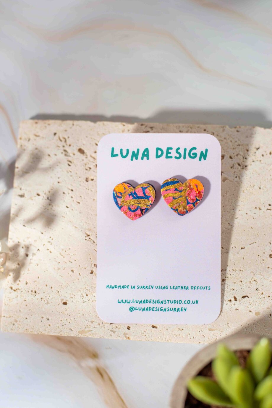 A pair of heart shaped earrings with orange gold and green accents displayed on a Luna Design branded card against a neutral stone background.