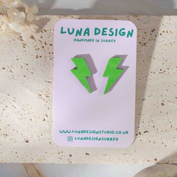 A pair of bright green lightning bolt-shaped earrings displayed on a Luna Design Studio card. The card features the brand name "Luna Design" in bold green text with "Handmade in Surrey" written below. The earrings are set on a textured stone surface with dried flowers and a small potted plant in the background.