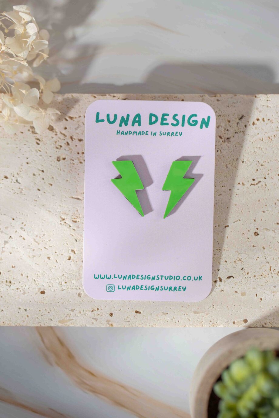 A pair of bright green lightning bolt-shaped earrings displayed on a Luna Design Studio card. The card features the brand name "Luna Design" in bold green text with "Handmade in Surrey" written below. The earrings are set on a textured stone surface with dried flowers and a small potted plant in the background.