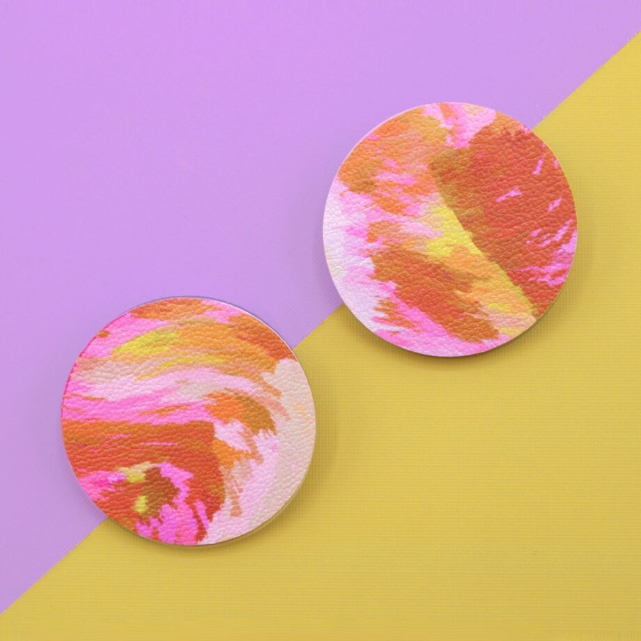 Flat lay of two round stud earrings featuring a pink, yellow and brown painterly abstract sunset design