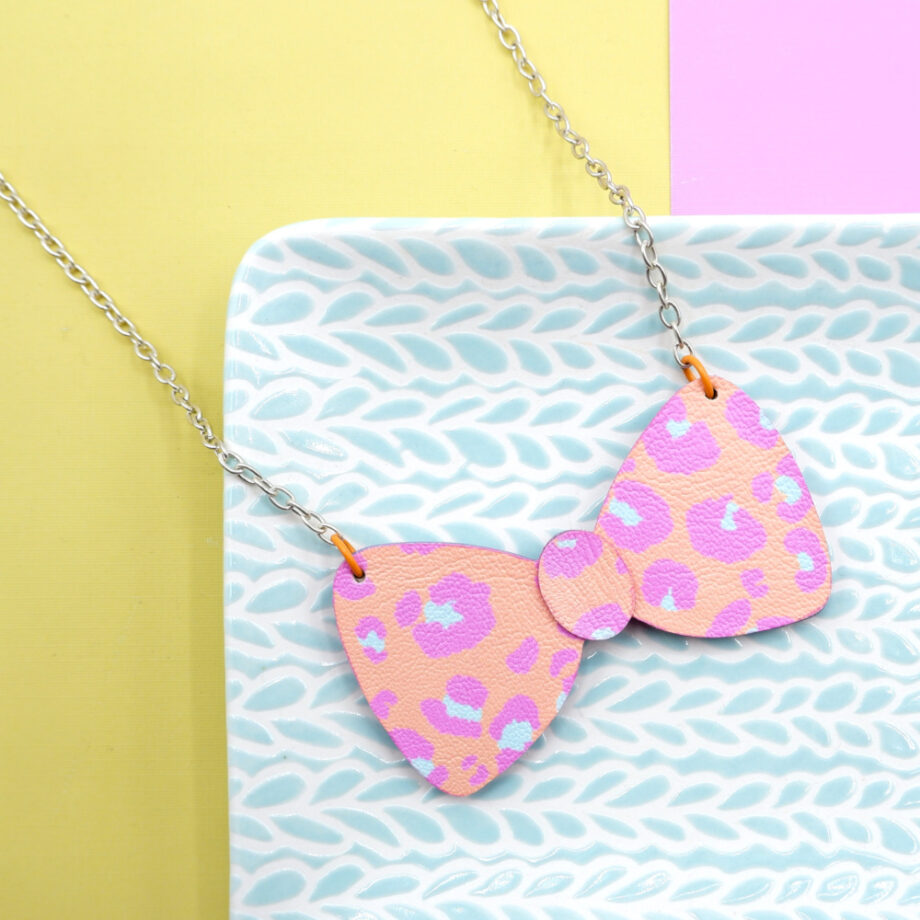 Quirky peach and pink leopard print necklace in the shape of a bow tie, with pale blue accents. Placed on pale blue textured decorative platter