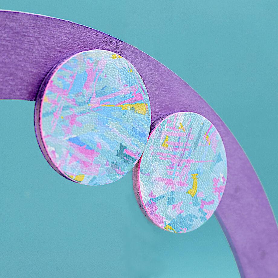 Close-up photo of the slightly side on view on a pair of circular earrings. The earrings are predomanently pale blue with flecks of pink, yellow and other shades of blue. Earrings shown displayed on a purple painted semi-circular stand