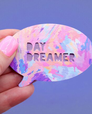 Handmade brooch with the text Day Dreamer cut from the surface of the painterly patterned faux leather. The metallic purple painted plywood base to the brooch shows throw