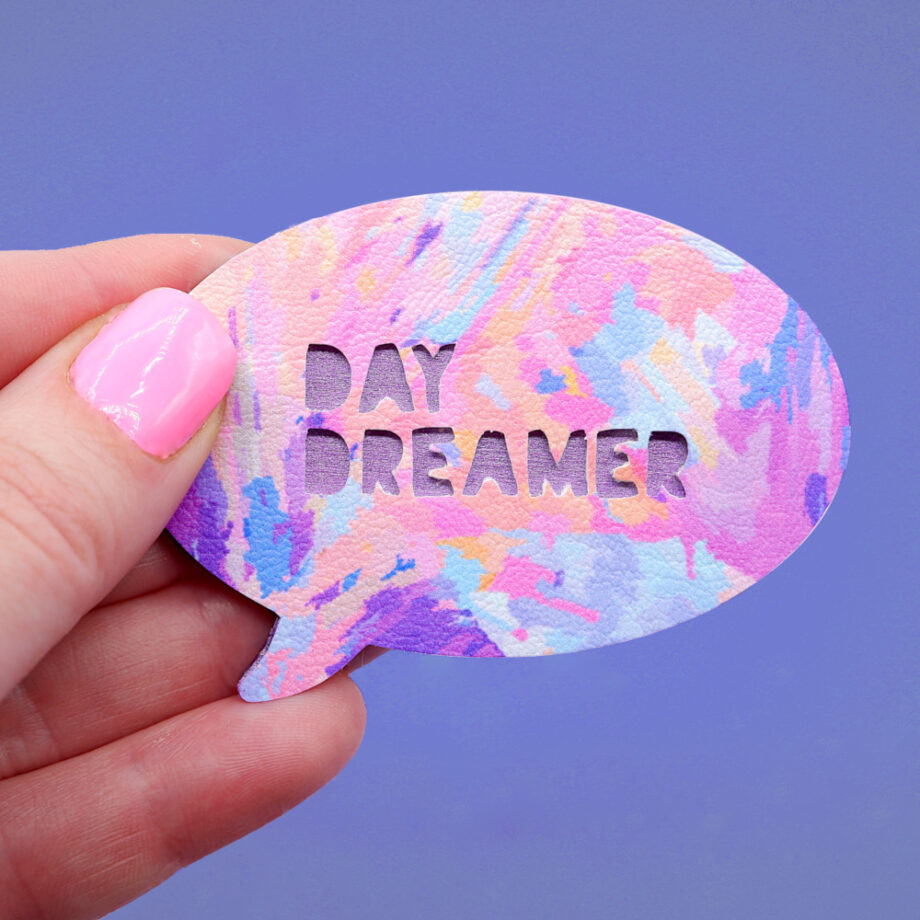 Handmade brooch with the text Day Dreamer cut from the surface of the painterly patterned faux leather. The metallic purple painted plywood base to the brooch shows throw