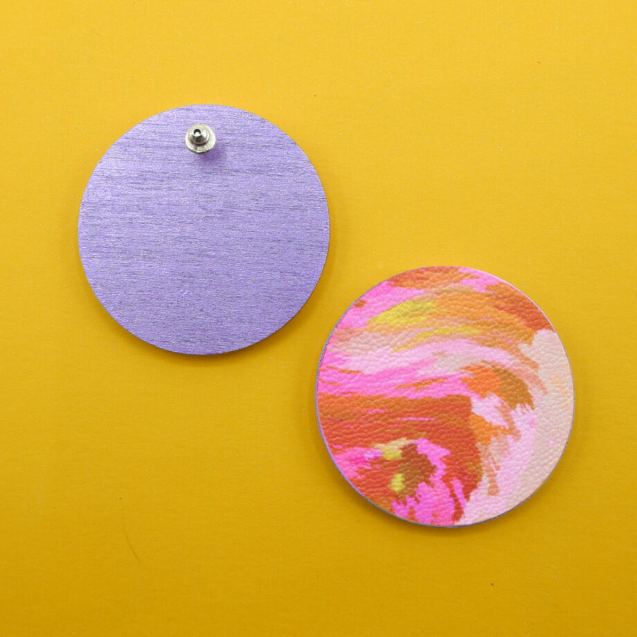 Close-up of circular stud earrings, showing a solid purple back and a vibrant, abstract patterned front with a 70's retro vibe, in pinks, yellows and brown