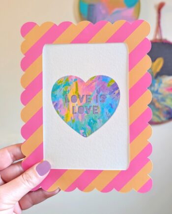A close-up shot shows a hand holding a framed rainbow heart mini art print. The print features a colorful heart with "LOVE IS LOVE" text, central to the design. A pink and peach striped, scalloped frame surrounds the artwork. In the blurred background, hand-painted abstract circular wall art hangs on a white wall.