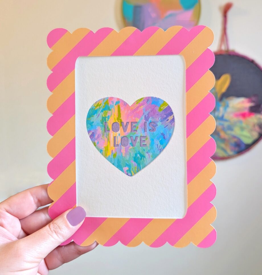 A close-up shot shows a hand holding a framed rainbow heart mini art print. The print features a colorful heart with "LOVE IS LOVE" text, central to the design. A pink and peach striped, scalloped frame surrounds the artwork. In the blurred background, hand-painted abstract circular wall art hangs on a white wall.