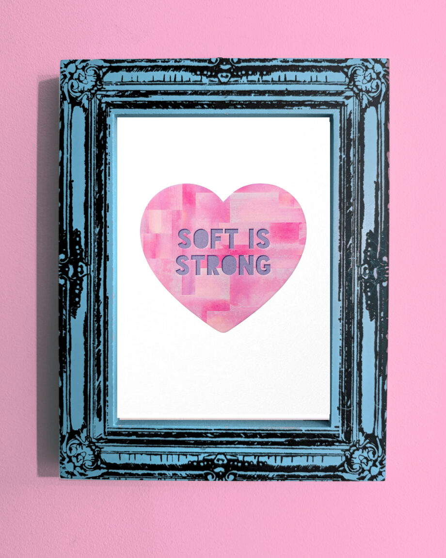 A framed art print displayed on a pink wall. The heart shape is filled with textured pink brush strokes and contains the phrase "SOFT IS STRONG". The ornate black frame has a distressed turquoise finish, adding contrast to the soft design.
