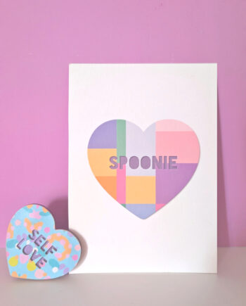 Pastel geometric heart art print with the word “Spoonie” on a white card, styled on a small shelf against a pink background.