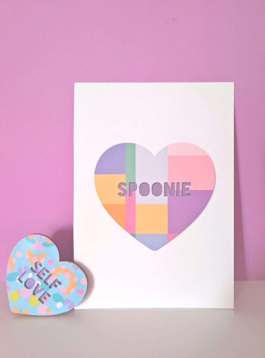 Pastel geometric heart art print with the word “Spoonie” on a white card, styled on a small shelf against a pink background.