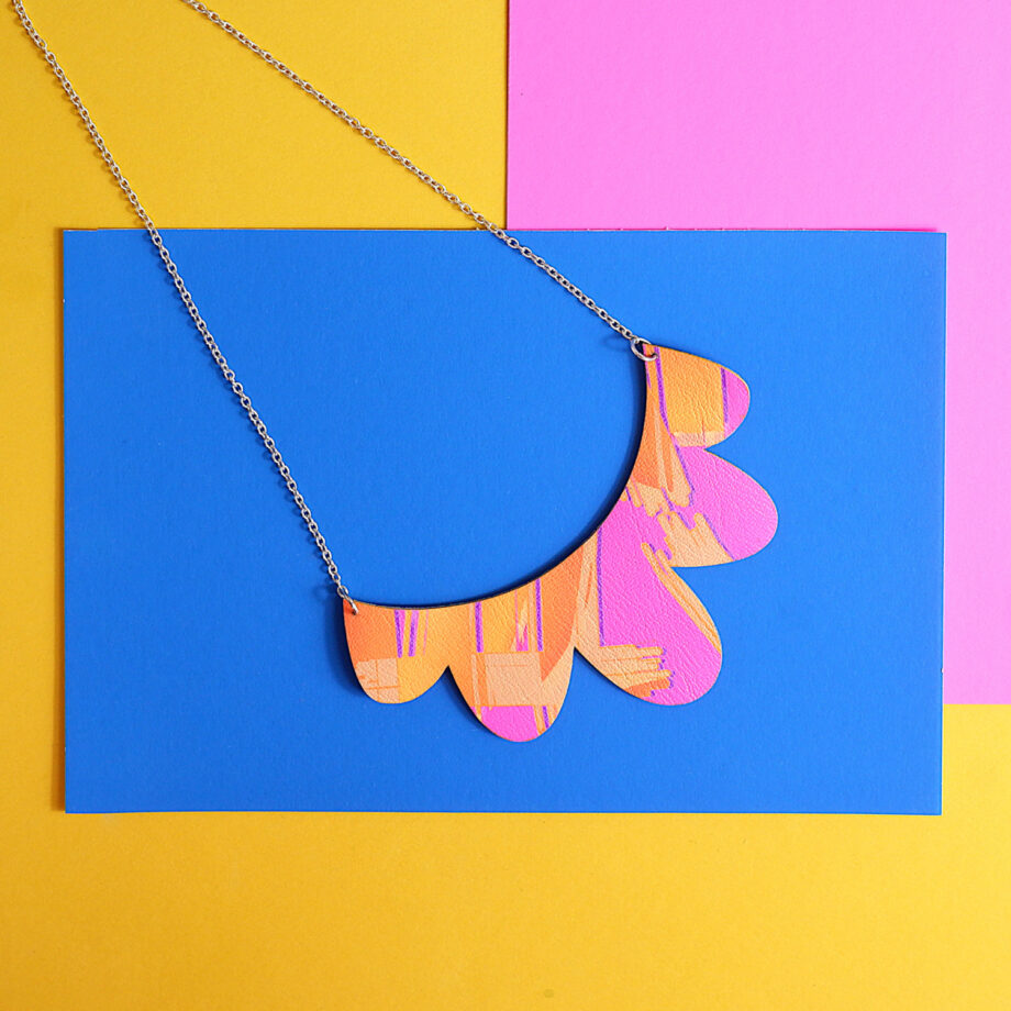 Query successful A vibrant, handmade statement necklace featuring a scalloped design. The necklace is crafted from faux leather, which is printed with a unique, original abstract pattern in shades of pink and orange. It hangs from a delicate silver-colored chain. The necklace is photographed on a background of bright blue, pink, and yellow.