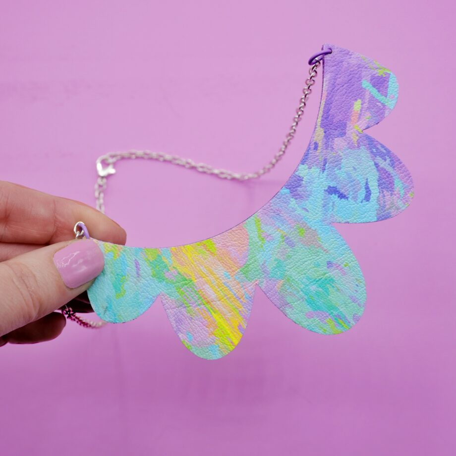 Handmade scalloped statement necklace made of faux leather with an abstract pastel rainbow print. The unique, lightweight necklace is displayed on a light blue patterned tray, featuring shades of lavender, mint green, and pale yellow, and is attached to a delicate silver chain.