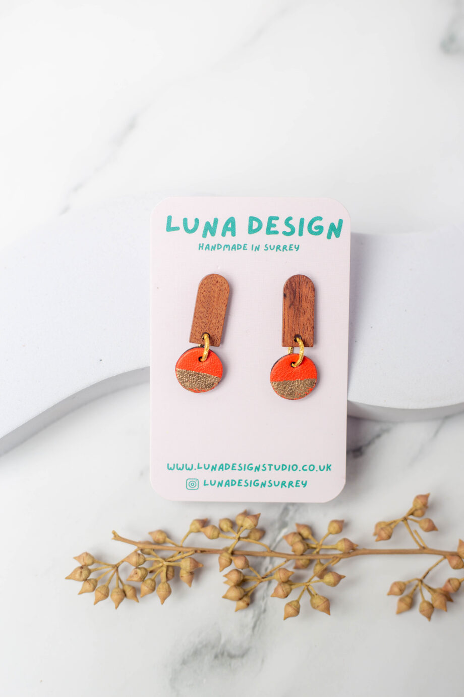 A pair of handmade wooden earrings with orange and gold half-moon drops, displayed on a "Luna Design" card. The earrings are photographed on a white marble surface with a sprig of dried foliage for decoration.